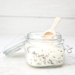 white bath salts with herbs in clear glass jar with clamp lid and wood scoop